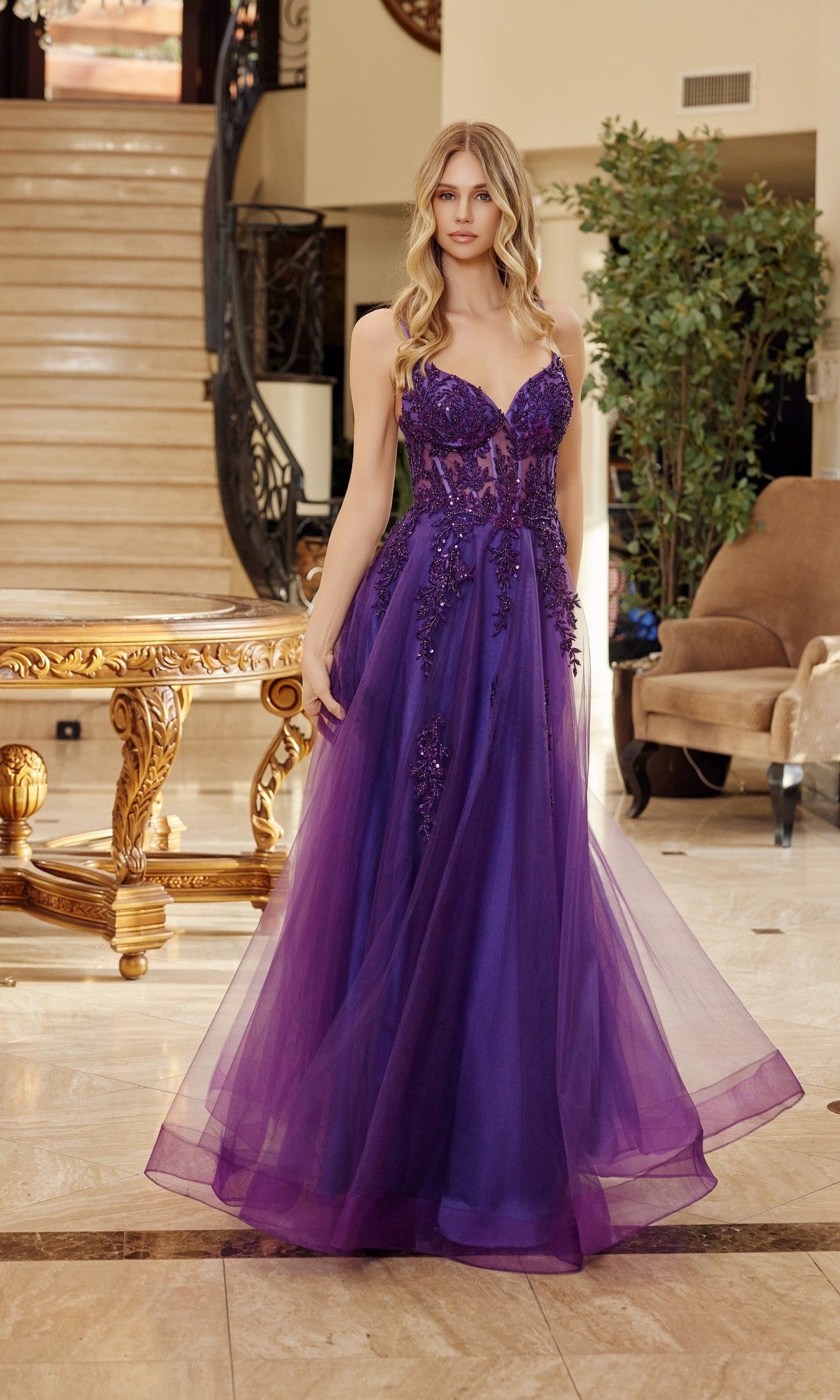 A woman stands indoors wearing the Nox Anabel Q1778 long prom dress, featuring tulle, floral embellishments, and a sheer embroidered corset bodice. Marble stairs and ornate furniture appear in the background.